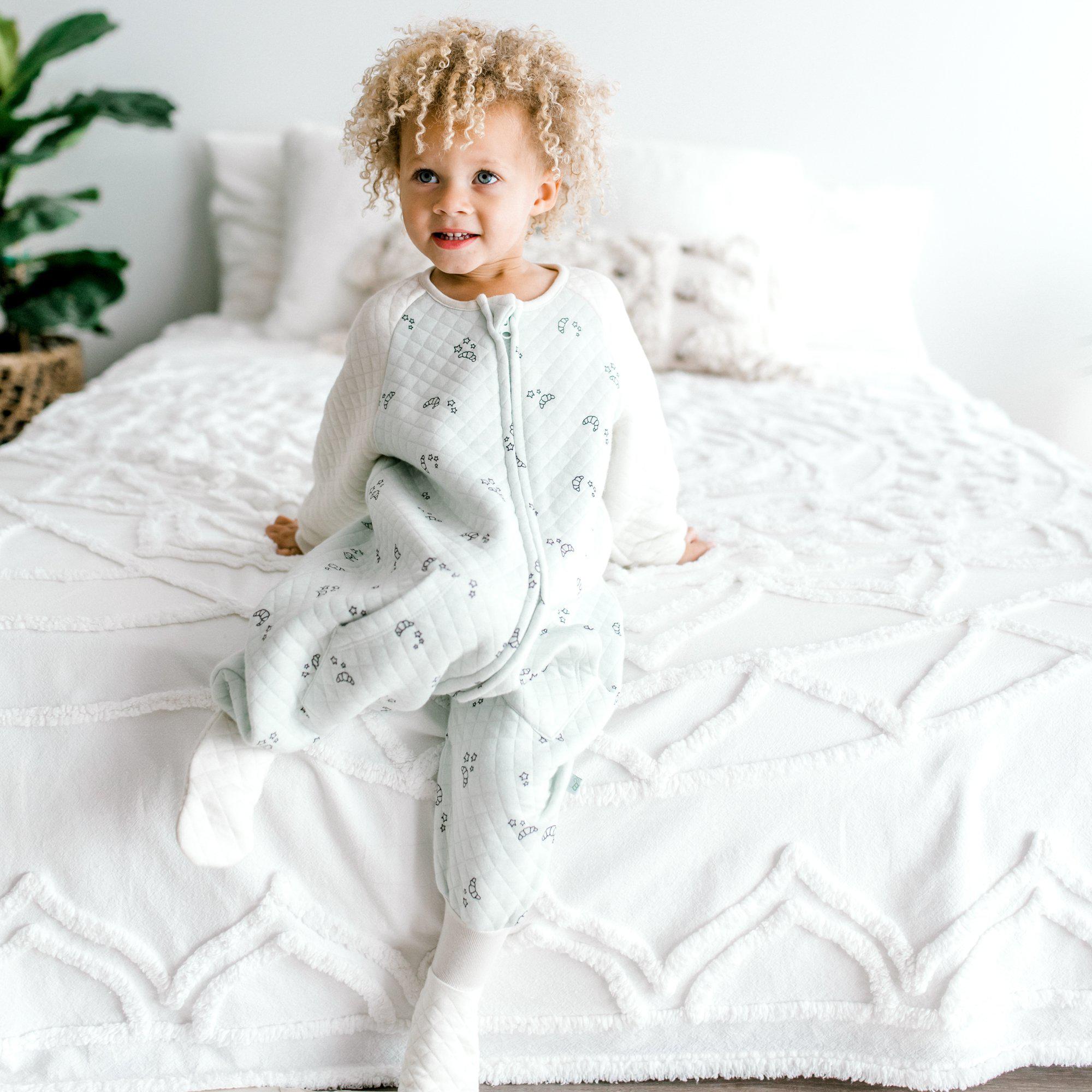 Full view of Baby wearing Sleep Sack With Sleeves Tealbee Dreamsie while sitting on the bed Croissant 0.8 TOG available from sizes 12m - 4T